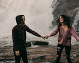 A couple laughing together holds hands in front of a large, powerful waterfall. The man is wearing a dark shirt with long sleeves, and the woman is in a light pink sweater with ripped jeans. Their expressions convey happiness and enjoyment.