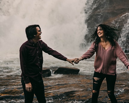 A couple laughing together holds hands in front of a large, powerful waterfall. The man is wearing a dark shirt with long sleeves, and the woman is in a light pink sweater with ripped jeans. Their expressions convey happiness and enjoyment.