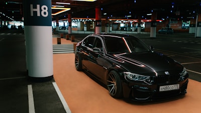 A sleek, black BMW car is parked in a dimly lit underground parking lot. The car is positioned near a column labeled H8. The setting has a modern and somewhat industrial feel with various overhead lights and another car visible in the background.