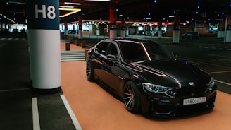 A sleek, black BMW car is parked in a dimly lit underground parking lot. The car is positioned near a column labeled H8. The setting has a modern and somewhat industrial feel with various overhead lights and another car visible in the background.
