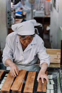 An elderly person dressed in a white chef's uniform is working with wooden slabs on a glass surface. There are various objects and containers in the blurred background, indicating a workplace setting, possibly a kitchen or workshop.