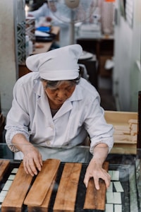 An elderly person dressed in a white chef's uniform is working with wooden slabs on a glass surface. There are various objects and containers in the blurred background, indicating a workplace setting, possibly a kitchen or workshop.
