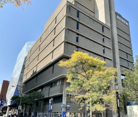 A large, modern multi-story building with a concrete facade is surrounded by trees with green and yellow leaves. Another glass building is partially visible in the background. Signs with the letter 'R' are displayed outside the building.