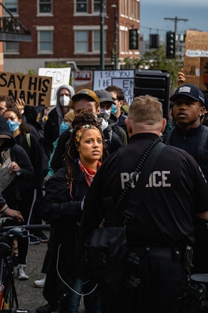 A group of people are gathered in a protest. Several hold signs with messages, including one with 'Say His Name.' A police officer, facing the crowd, stands in the foreground. The protesters appear serious and engaged, with some wearing masks.