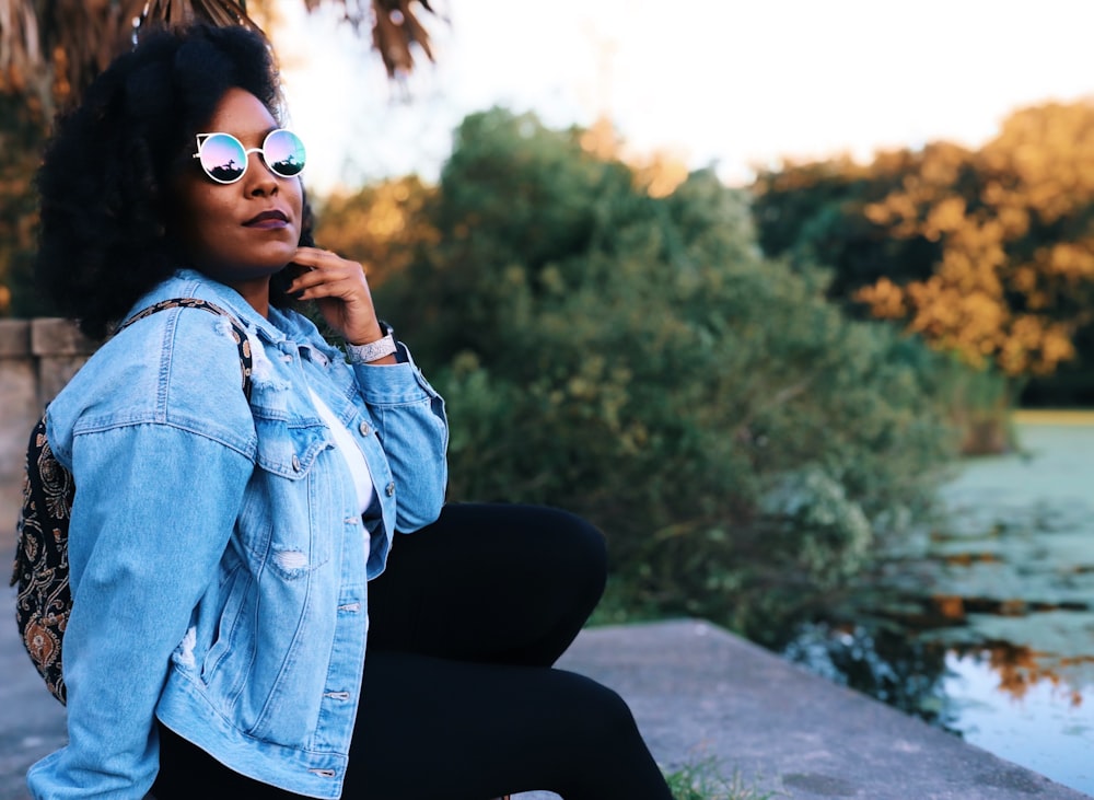 Femme en veste en jean bleue et pantalon noir assise sur un trottoir en béton gris pendant la journée