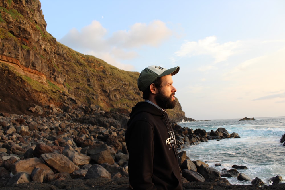 man in black jacket and gray cap standing on rocky shore during daytime