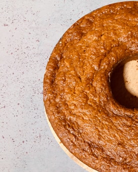 A golden-brown bundt cake with a textured surface is placed on a light speckled background. The cake has a smooth, uniform color and appears freshly baked, with a shine indicating a moist crumb.