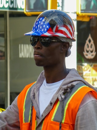 A person wears a construction helmet with an American flag design, along with sunglasses and a high-visibility orange safety vest over a grey hoodie.
