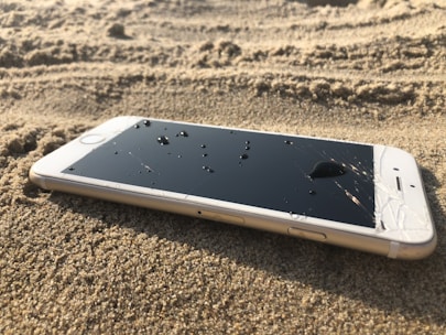 A smartphone lies face up on sandy ground, with water droplets on its cracked screen. The device appears to be slightly dusted with sand, suggesting it may have been dropped or left on the beach.