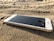 A smartphone lies face up on sandy ground, with water droplets on its cracked screen. The device appears to be slightly dusted with sand, suggesting it may have been dropped or left on the beach.