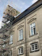 A large building with classic architectural features, including arched window frames and ornate detailing, is under renovation. Scaffolding is erected along the side, reaching up to the roof. The building has a muted, aged facade with multiple windows, some of which are open.