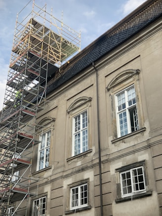 A large building with classic architectural features, including arched window frames and ornate detailing, is under renovation. Scaffolding is erected along the side, reaching up to the roof. The building has a muted, aged facade with multiple windows, some of which are open.