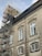A large building with classic architectural features, including arched window frames and ornate detailing, is under renovation. Scaffolding is erected along the side, reaching up to the roof. The building has a muted, aged facade with multiple windows, some of which are open.