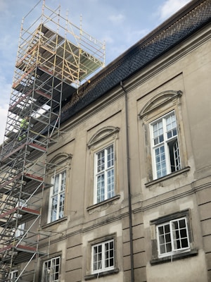 A large building with classic architectural features, including arched window frames and ornate detailing, is under renovation. Scaffolding is erected along the side, reaching up to the roof. The building has a muted, aged facade with multiple windows, some of which are open.