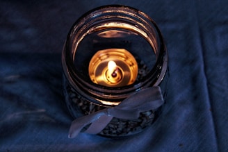 A lit candle is placed inside a transparent jar filled with small pebbles, and the jar is tied with a white ribbon. The jar is set on a textured blue surface.