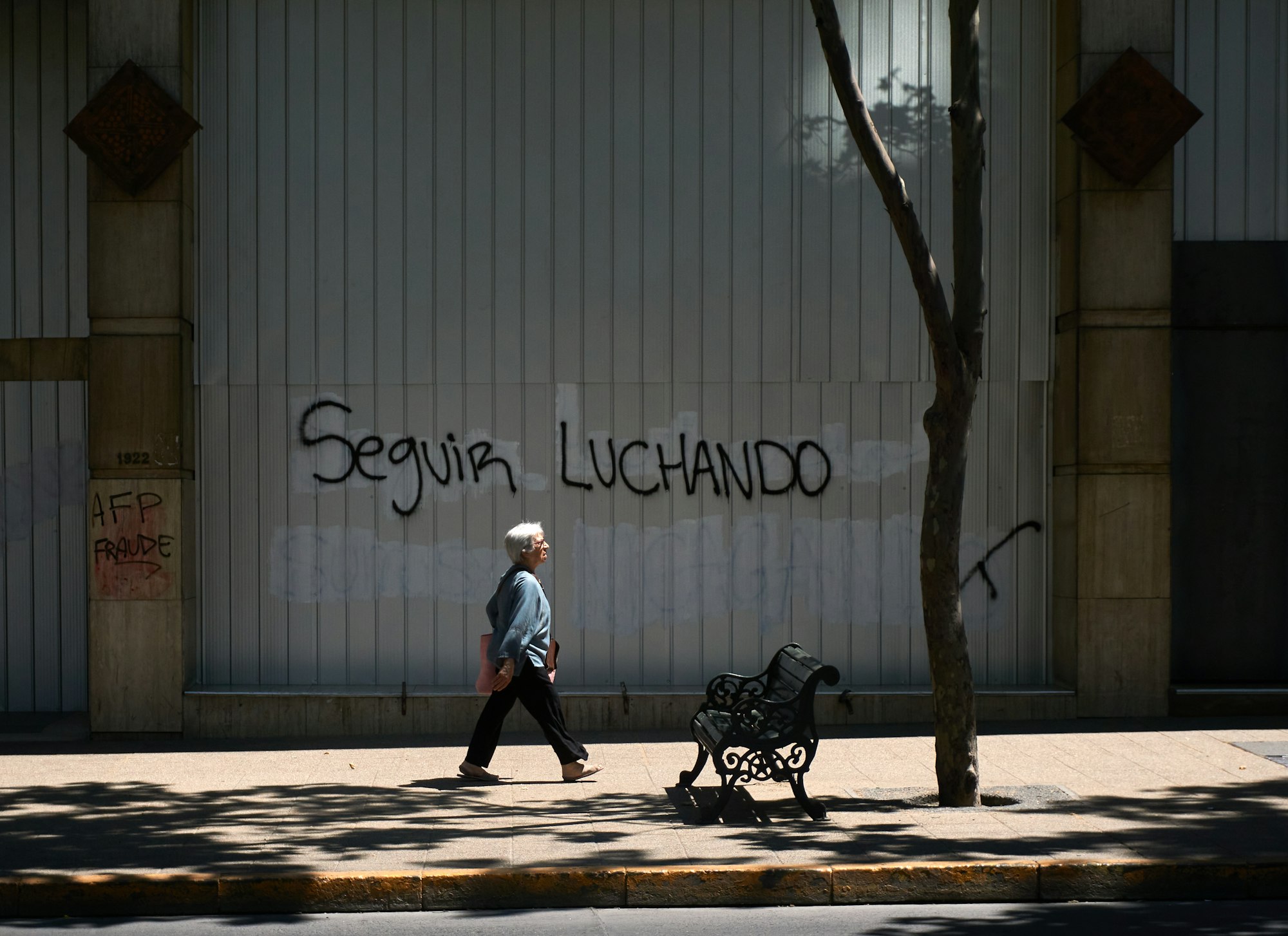 A person walks in front of grafitti written over a whitewashed wall, reading "Seguir Luchando" ("Keep Fighting")