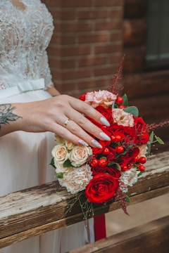 A close-up of a bridal bouquet featuring vibrant red roses, peach and white flowers, accented with red berries. A hand with a wedding ring and manicured nails rests gently on the bouquet. The background includes a wooden railing and a brick wall.