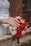 A close-up of a bridal bouquet featuring vibrant red roses, peach and white flowers, accented with red berries. A hand with a wedding ring and manicured nails rests gently on the bouquet. The background includes a wooden railing and a brick wall.