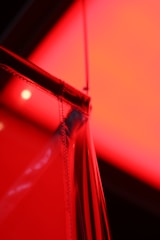 A close-up view of a translucent material in front of a bright, red light source. The material reflects and refracts the light, creating an abstract pattern. The scene has a smooth texture and glossy finish, with light playing off the edges, creating an intricate, artistic image.