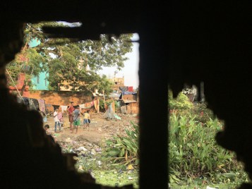 A group of children and a few adults are gathered in an open area filled with garbage and various debris. The setting appears to be a makeshift community or slum, with makeshift tents and structures constructed from tarps and cloth. Laundry is seen hanging on lines, and greenery with red flowers is present on one side. The image is framed by a dark, jagged border, possibly through a window or broken wall.