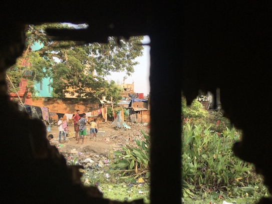 A group of children and a few adults are gathered in an open area filled with garbage and various debris. The setting appears to be a makeshift community or slum, with makeshift tents and structures constructed from tarps and cloth. Laundry is seen hanging on lines, and greenery with red flowers is present on one side. The image is framed by a dark, jagged border, possibly through a window or broken wall.