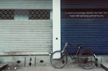 A closed storefront with two metal shutters, one painted gray and the other blue. A black bicycle is parked against the blue shutter. The blue shutter has a sign with text about privileges and banking. The ground is a dull, patterned surface.