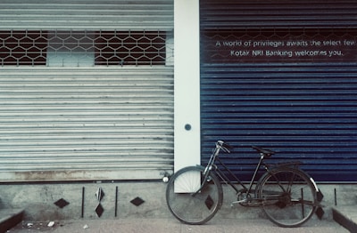 A closed storefront with two metal shutters, one painted gray and the other blue. A black bicycle is parked against the blue shutter. The blue shutter has a sign with text about privileges and banking. The ground is a dull, patterned surface.