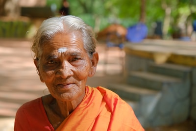 An elderly person with white hair and traditional clothing is sitting outdoors, surrounded by greenery. Soft sunlight brightens the scene, highlighting their serene expression.