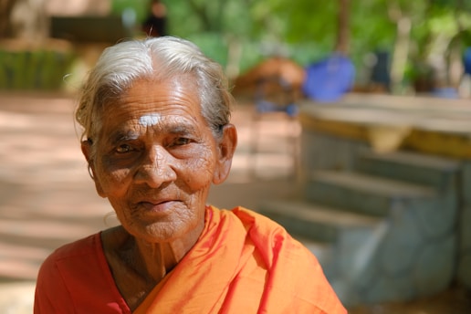 An elderly person with white hair and traditional clothing is sitting outdoors, surrounded by greenery. Soft sunlight brightens the scene, highlighting their serene expression.