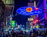 A bustling street at night filled with a variety of neon signs advertising places such as clubs, restaurants, and hotels. The street is crowded with people, showcasing a vibrant nightlife scene. The signs are prominently lit in colors like blue, green, red, and yellow, creating a lively and energetic atmosphere.