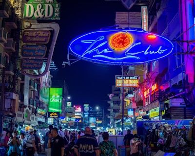 A bustling street at night filled with a variety of neon signs advertising places such as clubs, restaurants, and hotels. The street is crowded with people, showcasing a vibrant nightlife scene. The signs are prominently lit in colors like blue, green, red, and yellow, creating a lively and energetic atmosphere.