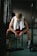 A person sits on a bench in a dimly lit gym, hunched over with elbows resting on their knees. They wear a white sleeveless shirt with a towel draped around their neck, black shorts, white socks, and athletic shoes. A barbell with weights is on the floor in front of them. The mood suggests fatigue or contemplation, with the person's gaze directed downward.