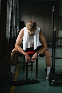 A person sits on a bench in a dimly lit gym, hunched over with elbows resting on their knees. They wear a white sleeveless shirt with a towel draped around their neck, black shorts, white socks, and athletic shoes. A barbell with weights is on the floor in front of them. The mood suggests fatigue or contemplation, with the person's gaze directed downward.