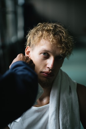 A person with curly blond hair looks directly at the camera. They have a white towel draped over their shoulders and wear a white tank top. A blurred hand in the foreground is gently wiping their face, which appears slightly sweaty. The background is out of focus, suggesting a dimly lit indoor environment.