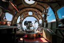 The interior of a vintage aircraft cockpit with a single pilot seat at the center, surrounded by various controls, levers, and instruments. The large windows offer a view of the sky and mountains, creating a contrast between the mechanical interior and the natural landscape. Sunlight streams through the windows, illuminating the metallic structure.