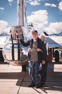 A friendly older couple smiling beside a classic Cadillac car in a sunny park setting.