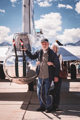 A friendly older couple smiling beside a classic Cadillac car in a sunny park setting.