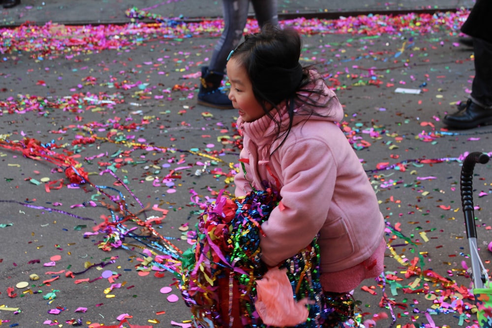 girl in pink jacket and blue pants sitting on ground with pink leaves