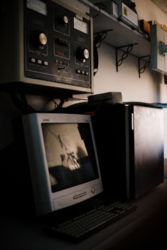 A vintage computer setup featuring an old CRT monitor and traditional keyboard is placed on a desk. Above the desk, there is industrial equipment with large knobs and dials, suggesting an older control system or machinery interface. Shelving with various equipment and boxes is visible in the background.