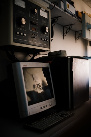 A vintage computer setup featuring an old CRT monitor and traditional keyboard is placed on a desk. Above the desk, there is industrial equipment with large knobs and dials, suggesting an older control system or machinery interface. Shelving with various equipment and boxes is visible in the background.