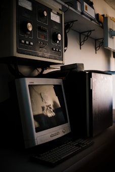 A vintage computer setup featuring an old CRT monitor and traditional keyboard is placed on a desk. Above the desk, there is industrial equipment with large knobs and dials, suggesting an older control system or machinery interface. Shelving with various equipment and boxes is visible in the background.