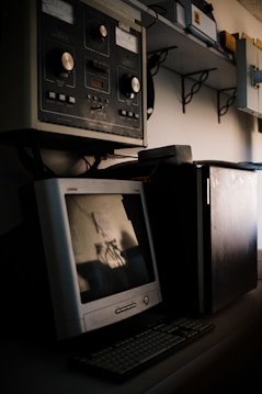 A vintage computer setup featuring an old CRT monitor and traditional keyboard is placed on a desk. Above the desk, there is industrial equipment with large knobs and dials, suggesting an older control system or machinery interface. Shelving with various equipment and boxes is visible in the background.