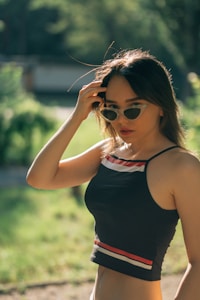 A person is wearing sunglasses and a sleeveless top with stripes, standing outdoors with sunlight creating a warm, natural glow. The background consists of blurred trees and greenery.