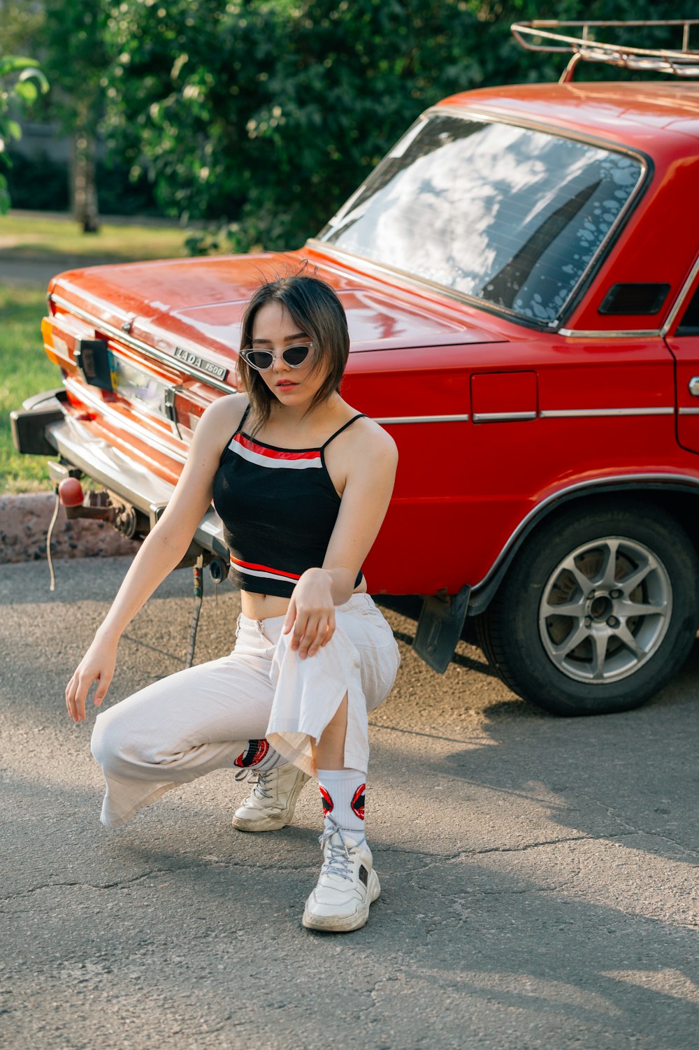 Woman in black tank top and white pants standing beside red car