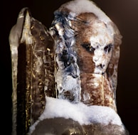 A detailed sculpture of a face partially encased in ice, showing intricate features like deep-set eyes. The sculpture is dusted with snow, adding a cold and frozen texture to its surface. The ice surrounding it is clear in places, revealing the underlying details.