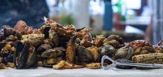 A variety of seafood and accompaniments displayed on a table, including crabs, clams, mussels, corn on the cob, and sausages. The ingredients are cooked and mixed together, creating a rustic and hearty presentation.
