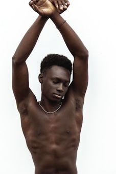 A person with short curly hair is posing with arms crossed above the head, eyes are closed indicating a calm or meditative state. The background is plain and white, accentuating the subject's skin tone.