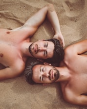 Two shirtless men with short hair lie on sandy ground, with their heads close together. They gaze directly at the camera with relaxed expressions. The sand surrounding them adds a warm and natural backdrop.