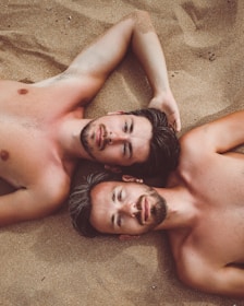 Two shirtless men with short hair lie on sandy ground, with their heads close together. They gaze directly at the camera with relaxed expressions. The sand surrounding them adds a warm and natural backdrop.
