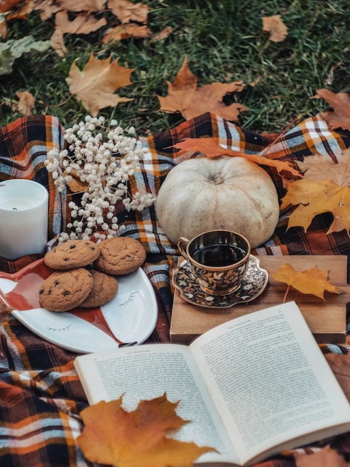A cozy autumn-themed picnic setup features a plaid blanket surrounded by fallen leaves. At the center of the scene, a plate with chocolate chip cookies, a decorative cup of black coffee, and an open book create an inviting atmosphere. A small pumpkin and a bouquet of white flowers add seasonal charm, while a lit candle enhances the warm ambiance.
